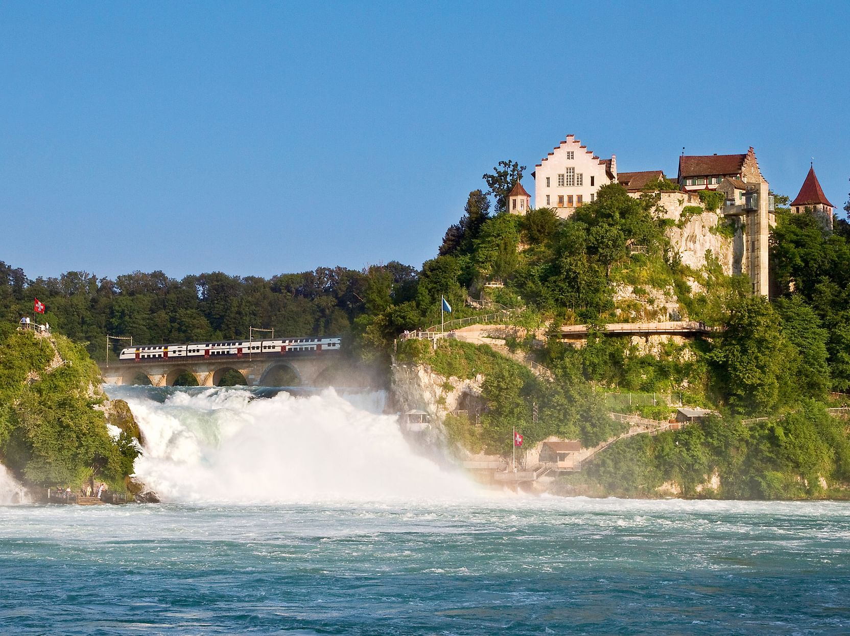 Rheinfall-Wasserfall mit weißem kaskadierendem Wasser und Nebel. Das historische Schloss Laufen thront auf grünen Klippen darüber.