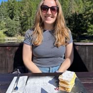 Frau mit Sonnenbrille an Tisch mit Cremeschnitte auf Schieferplatte im Restaurant zum See in Grächen, bewaldete Berge im Hintergrund.