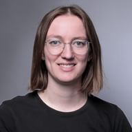 Porträt von Jael Loser mit schulterbraunem Haar, runder Brille und schwarzem Shirt, lächelnd in die Kamera vor grauem Hintergrund.