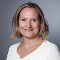 Portrait professionnel de Tanja Joss avec des cheveux blonds mi-longs, portant un haut blanc et un collier, souriant sur fond gris.