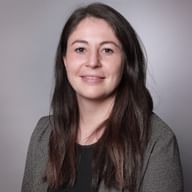 Professional headshot of Cindy Mayor with long brown hair, wearing a gray blazer over a dark top, smiling at the camera against a gray background.
