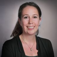 Professionelles Porträt von Romaine Bayard, einer Frau mit braunen Haaren in dunklem Blazer und Halskette, lächelnd vor grauem Hintergrund.