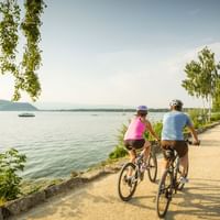 Zwei Radfahrer auf einem gepflasterten Weg am Seeufer der Mittelland-Route. Die Frau trägt Rosa, der Mann Blau. See und Berge im Hintergrund.