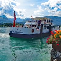 Bateau de passagers blanc nommé Salzkammergut sur le lac Wolfgangsee avec drapeau autrichien, montagnes en arrière-plan, fleurs colorées au premier plan.