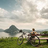 Eine Frau sitzt auf einer Bank und macht Pause mit Aussicht auf den Lago di Lugano und den San Salvatore.