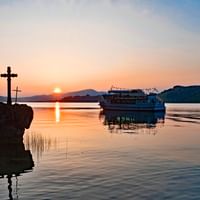Vue du coucher de soleil sur le lac Mondsee avec un bateau de passagers ancré dans l'eau calme. Une croix en pierre se dresse au premier plan.
