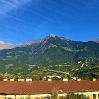 Vue panoramique du Meraner Oberland avec des sommets alpins enneigés, des montagnes boisées vertes et des bâtiments résidentiels jaunes.