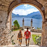 Zwei Touristen gehen durch Steinbogen in Morcote mit Blick auf Luganer See, Bergen und blauem Himmel im Hintergrund, Tessin, Schweiz.