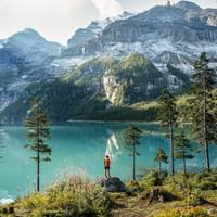 Hiker at turquoise Oeschinensee lake Hiker in red jacket standing on rocky outcrop overlooking brilliant turquoise Oeschinensee lake surrounded by snow-capped Alpine peaks and forest.