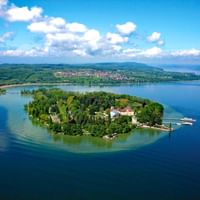 Vue aérienne de l'île de Mainau dans le lac de Constance avec des forêts vertes denses, des bâtiments historiques et un petit port.