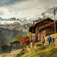 Zwei Wanderer gehen zu traditionellen Holz-Berghütten mit Schweizer Flagge, umgeben von schneebedeckten Alpengipfeln nahe Saas Fee.