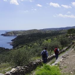 Zwei Wanderer mit Rucksäcken auf einem steingemauerten Weg mit Blick auf die Mittelmeerküste bei Cap de Creus, mit grüner Vegetation und blauem Meer.