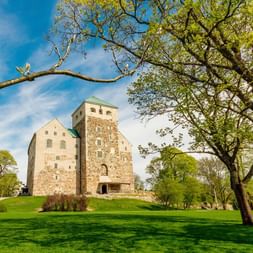 Burg Turku mit Steinmauern und grünem Kupferdach, umgeben von saftig grünem Rasen und belaubten Bäumen unter blauem Himmel mit weißen Wolken.
