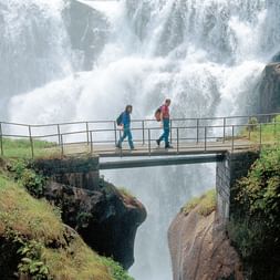 Zwei Personen auf einer Brücke und im Hintergrund ein grosser Wasserfall. Via Suworow. Wanderferien mit Eurotrek.