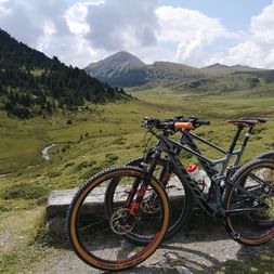 Vorne sind 2 Mountainbikes angelehnt an einen Holzblock vor einer Landschaft aus grünen Bergen und Wiesen.