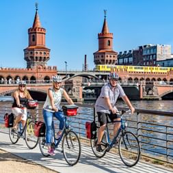Trois cyclistes avec casques debout avec leurs vélos sur une promenade au bord de l'eau devant le pont historique Oberbaum de Berlin.