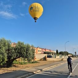 Radfahrer auf einer Straße in Nordkatalonien mit einem gelben Heißluftballon am blauen Himmel über Wohngebäuden.