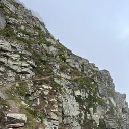 Wanderin auf schmalem Pfad entlang steiler Felswand am Alpenpässeweg. Der Weg führt über eine dramatische Bergflanke mit Felsschichten.