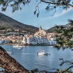 Mediterranes Küstendorf mit weißen Häusern und Kirche durch Pinienzweige gesehen. Boote in blauer Bucht, Berge im Hintergrund.