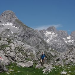 Wanderer in blauem Hemd geht durch felsiges Gelände am Peja Pass mit dramatischen Kalksteingipfeln und Schneeflecken unter blauem Himmel.