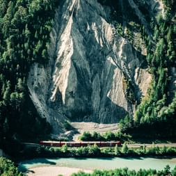 Red train traveling along turquoise river beneath dramatic gray rock formations surrounded by dense green forest in Graubünden, Switzerland.