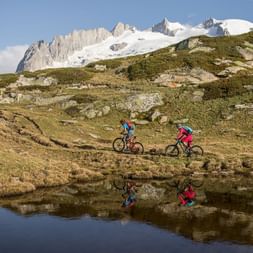 Zwei Mountaikbiker fahren über eine Wiese, hinter einem kleinen See und wundervollen, leicht mit Schnee bedekten Bergspitzen.