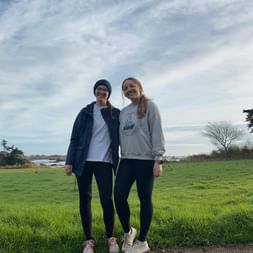 Zwei Frauen stehen auf grüner Wiese auf Île de Bréhat mit Küstenblick. Beide tragen Freizeitkleidung und lächeln in die Kamera.