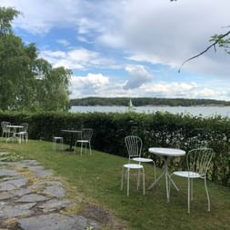Gemuetliche Sitzgelegenheit in Finnland am Meer. Im Vordergrund hat es weisse kleine Runde Tische mit weissen Stühlen auf einer Wiese. Im Hintergrund hat es Bäume, Hecken und das Meer.