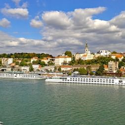Panoramablick auf Belgrad mit weißen Flusskreuzfahrtschiffen am Ufer und historischen Gebäuden mit Kirchturm auf dem Hügel.