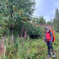 Wanderin Sara mit orangem Rucksack und roter Weste auf Weg umgeben von rosa Wildblumen, Vogelbeerbaum mit roten Beeren und Nadelbäumen.