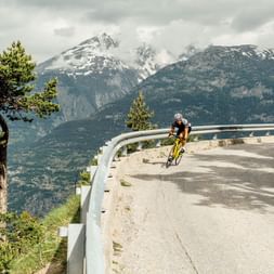 Cyclist coming out of a bend in a mountain landscape with snow-covered peaks.