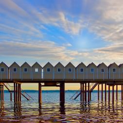 Reihe bunter Strandhütten auf Holzpier über dem Wasser in Helsingborg, Schweden. Dramatischer Himmel mit Sonnenstrahlen und Wasserreflexionen.
