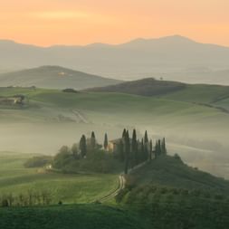Rolling green hills of Tuscany at sunrise with morning mist, featuring a hilltop villa surrounded by tall cypress trees and winding dirt road.