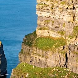 Hohe Kalksteinklippen in Westirland mit geschichteten Felsformationen und grünem Gras oben, neben einer hohen Felsnadel im blauen Ozean.