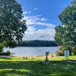 Ruhiger Seeblick in Bommersvik bei Stockholm mit grüner Wiese, großen Bäumen und Häusern am Ufer. Waldlinie über dem Wasser unter blauem Himmel.
