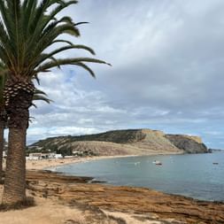 Palmengesäumte Promenade in Praia da Luz mit Blick auf Sandstrand, ruhige Bucht mit Booten und dramatische Klippe unter bewölktem Himmel.