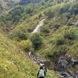 Zwei Wanderer mit Rucksäcken auf Bergpfad in der Ostschweiz. Ein Wasserfall fließt grüne Hänge hinab mit Felsklippen darüber.