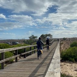 Zwei Radfahrer auf einem Holzsteg durch den Naturpark Ria Formosa in der Algarve. Grüne Vegetation und bewölkter Himmel sichtbar.