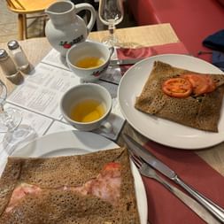 Zwei Buchweizen-Galettes auf weißen Tellern in einem Restaurant in Paimpol, eine mit Schinken und eine mit Tomaten, serviert mit Cidre in Schalen.