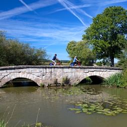 Zwei Radfahrer überqueren eine historische Steinbogenbrücke bei Ansbach. Die Brücke überspannt einen ruhigen Fluss mit Seerosen.