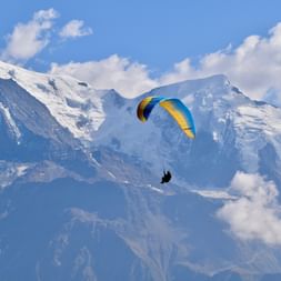 Gleitschirmflieger mit gelb-blauem Schirm vor schneebedeckten Mont Blanc Gipfeln unter blauem Himmel mit weißen Wolken.