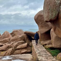 Person in blauer Jacke auf Steinweg zwischen massiven rosa Granitfelsen an der Küste von Ploumanach, Bretagne. Bewölkter Himmel und Meer im Hintergrund.