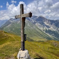 Hölzernes Gipfelkreuz auf grasigem Gipfel mit dramatischen Alpengipfeln im Hintergrund unter blauem Himmel mit weißen Wolken.