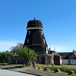 Schwarze Holzwindmühle mit weißen Fenstern auf Steinfundament in Simrishamn, umgeben von grünem Rasen und Bäumen unter blauem Himmel.