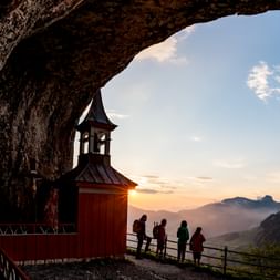 Rote Holzkapelle mit Glockenturm in einer Höhlenöffnung am Ebenalp. Besucher stehen auf Aussichtsplattform und beobachten Sonnenuntergang über Alpen.