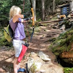 Frau mit blonden Haaren zielt mit Pfeil und Bogen auf eine Zielscheibe im Wald in Pfunds, Tirol. Sie trägt ein lila Shirt und steht auf felsigem Boden.