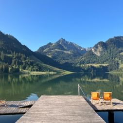 Wooden pier on Schwarzsee lake with two orange deck chairs, forested mountains reflected in calm water under clear blue sky near Bern.