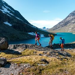 Drei Wanderer gehen über felsiges Gelände im Svartdalen Valley, Norwegen, mit einem türkisfarbenen Bergsee und schneebedeckten Gipfeln im Hintergrund.
