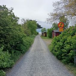 Schotterweg gesäumt von grüner Vegetation und roten Holzgebäuden. Ein Verkehrsverbotsschild ist rechts sichtbar.