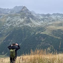 Wanderer mit grünem Rucksack fotografiert Alpengipfel von goldener Wiese bei Gruben. Bergkämme und bewaldete Täler im Hintergrund sichtbar.